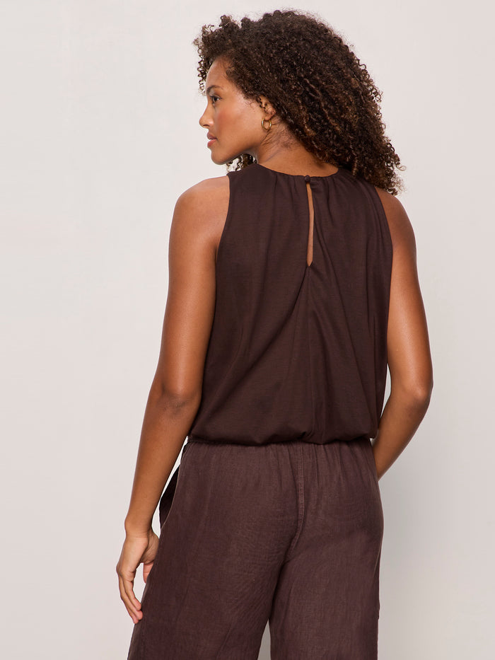 A woman with curly hair is seen from the back, wearing Sanctuary Clothing’s retreat bubble tank in rich soil—a sleeveless dark brown top with a keyhole—paired with matching loose brown pants against a plain light background.
