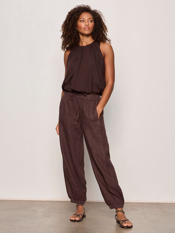 A woman with curly hair stands confidently in Sanctuary Clothing's retreat bubble tank in rich soil, paired with matching loose drawstring pants and sandals, against a plain light wall.