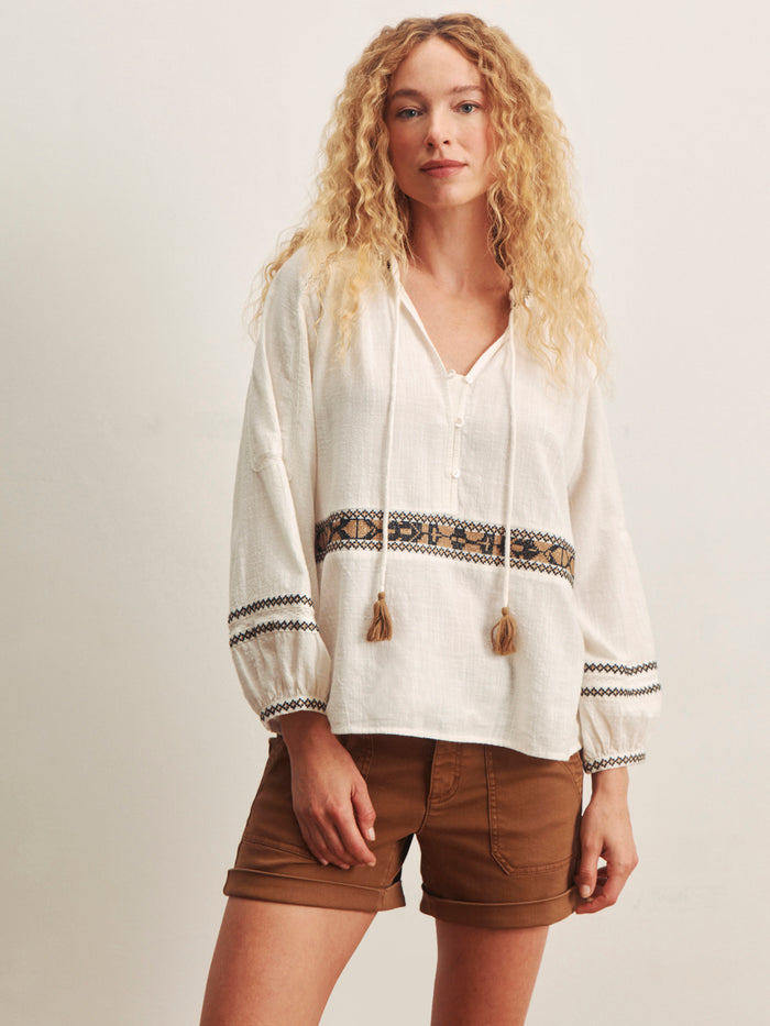 A woman with long curly blonde hair wears the Sanctuary Clothing retreat embroidered blouse in chalk, featuring geometric patterns and tassels, paired with brown shorts against a plain light background.