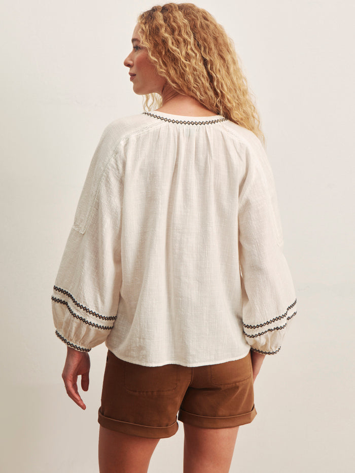 A woman with curly blonde hair wears the Sanctuary Clothing retreat embroidered blouse in chalk and brown shorts, standing against a plain background and looking over her shoulder.