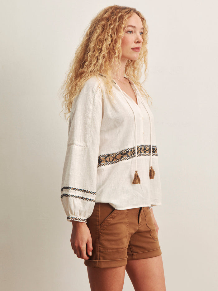 A woman with long curly blonde hair stands in profile, wearing the Sanctuary Clothing retreat embroidered blouse in chalk and brown shorts against a plain light background.