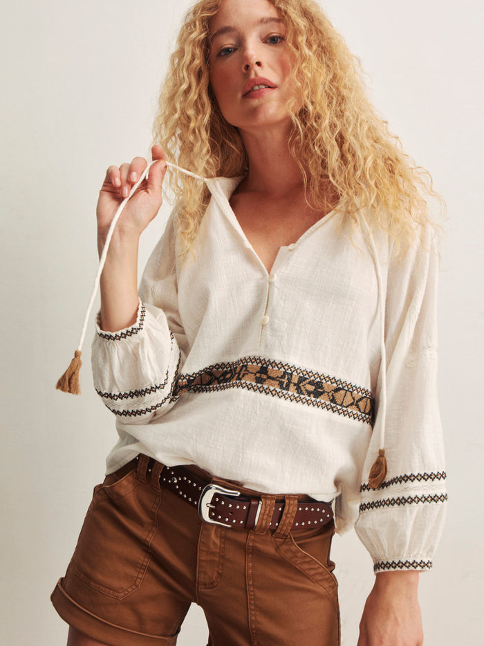 A woman with long curly blonde hair wears the Sanctuary Clothing retreat embroidered blouse in chalk, styled with brown shorts. She holds a tassel from the blouse and stands against a plain light background.
