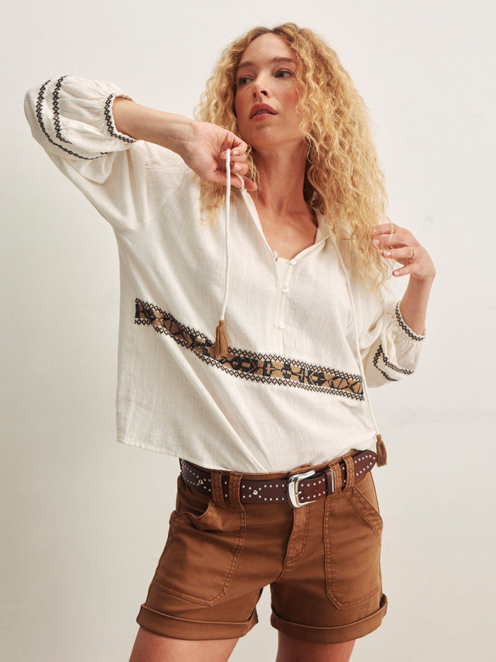 A woman with long curly blonde hair models the Sanctuary Clothing retreat embroidered blouse in chalk, featuring a loose fit with patterned trim, paired with high-waisted brown shorts against a plain light background.