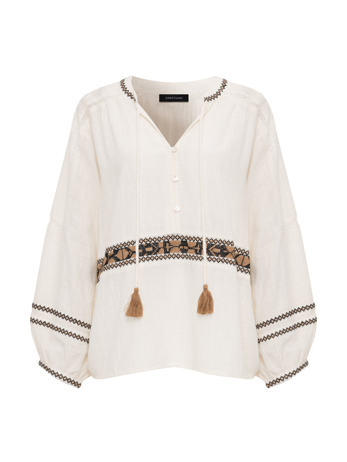 The retreat embroidered blouse chalk by Sanctuary Clothing is a cream long-sleeve top with a V-neck, front buttons, tassel ties, and black and tan embroidery on the sleeves, chest, and hem.