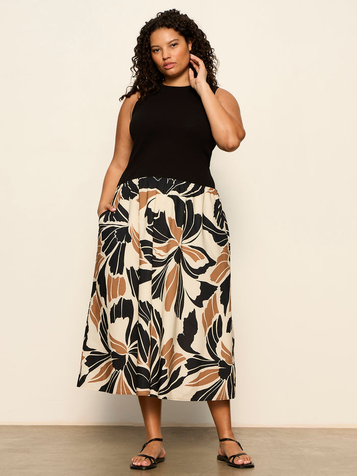 A woman stands against a light wall, wearing the Sanctuary Clothing sandbar mix media midi dress in lotus with black (extended sizing). The dress features bold beige, black, and brown floral patterns. She has curly hair, sandals, and one hand in her pocket.