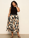 A woman stands against a light wall, wearing the Sanctuary Clothing sandbar mix media midi dress in lotus with black (extended sizing). The dress features bold beige, black, and brown floral patterns. She has curly hair, sandals, and one hand in her pocket.