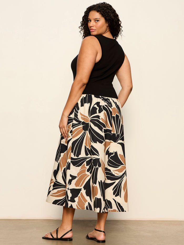 A woman with curly hair, wearing the Sanctuary Clothing sandbar mix media midi dress in lotus with black (extended sizing), stands on a light-colored floor facing away and looking over her shoulder.