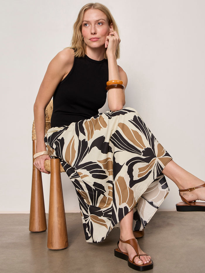 A woman with blonde hair sits on a woven chair, wearing the Sanctuary Clothing sandbar mix media midi dress in lotus w black, paired with brown sandals and a chunky brown bangle.