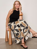 A woman with blonde hair sits on a woven chair, wearing the Sanctuary Clothing sandbar mix media midi dress in lotus w black, paired with brown sandals and a chunky brown bangle.
