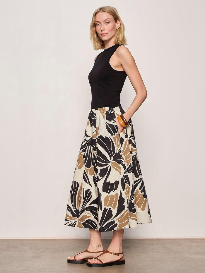 A woman with short blonde hair wears the Sanctuary Clothing sandbar mix media midi dress in lotus w black, featuring a sleeveless black top and a bold, abstract floral-print skirt. She stands sideways and pairs the look with flat sandals.