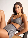 A woman with long, dark hair reclines on a cane chair, wearing the Sanctuary Clothing scoop front bra bikini top in navy. She gazes at the camera with a relaxed expression.