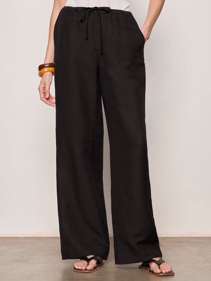 A person stands against a plain light background wearing Sanctuary Clothing shoreline linen pants in black, paired with a white sleeveless top, black flat sandals, and a brown bangle on their left wrist.