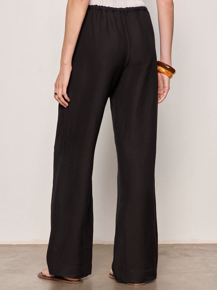 A person wearing Sanctuary Clothing shoreline linen pants in black and flat sandals stands on a gray floor, facing away from the camera, with a brown bangle on their right wrist.