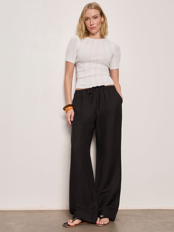 A woman with a neutral expression stands against a plain wall, hands in her pockets, wearing Sanctuary Clothing Shoreline Linen Pants in black, paired with a fitted white short-sleeve top, brown sandals, and a chunky brown bracelet.