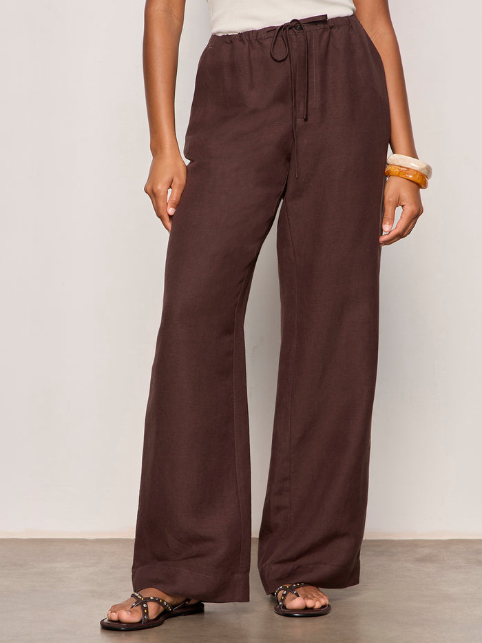 Someone wearing Sanctuary Clothing’s Shoreline Linen Pant in Rich Soil—loose, dark brown drawstring pants—paired with a white sleeveless top, black studded sandals, and chunky bangles, stands against a plain light background.