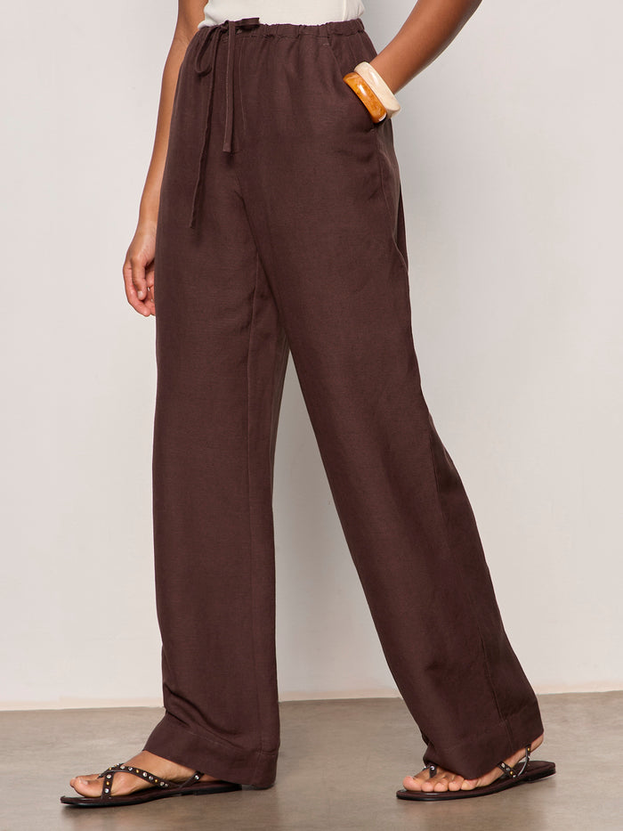 A person models Sanctuary Clothing's Shoreline Linen Pant in Rich Soil, paired with studded brown sandals and a tan clutch. The image features a plain light background and is cropped at the chest.