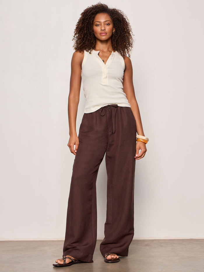 A woman with curly hair poses confidently against a plain background in Sanctuary Clothing’s shoreline linen pant in rich soil, paired with a sleeveless cream henley top, black sandals, and gold bangles.
