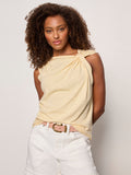 A woman with curly brown hair wears the Sanctuary Clothing "so twisted muscle tee banana," a sleeveless light yellow top with gathered shoulder detail, paired with white high-waisted shorts and a pink belt, against a plain background.