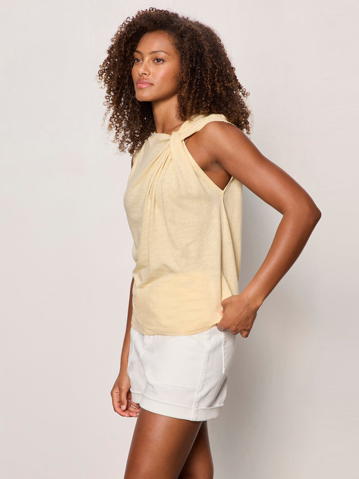 A woman with curly hair wears the Sanctuary Clothing "so twisted muscle tee banana," a sleeveless, knotted-shoulder yellow top, paired with white shorts and standing against a plain light background.