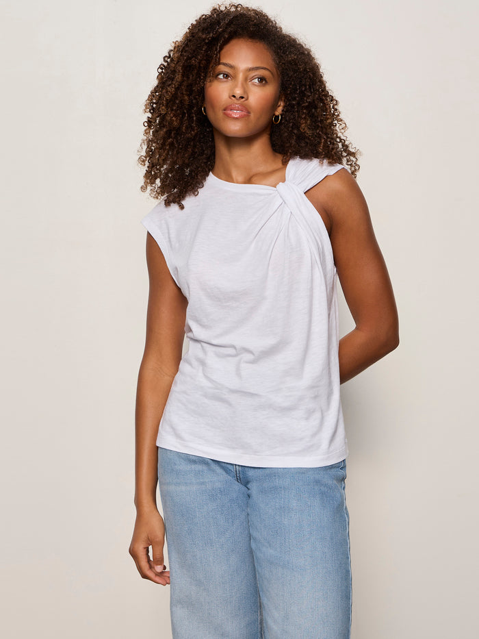 A woman with curly hair wears the Sanctuary Clothing So Twisted Muscle Tee in white, featuring a knotted shoulder detail, paired with light blue jeans and standing against an off-white background.