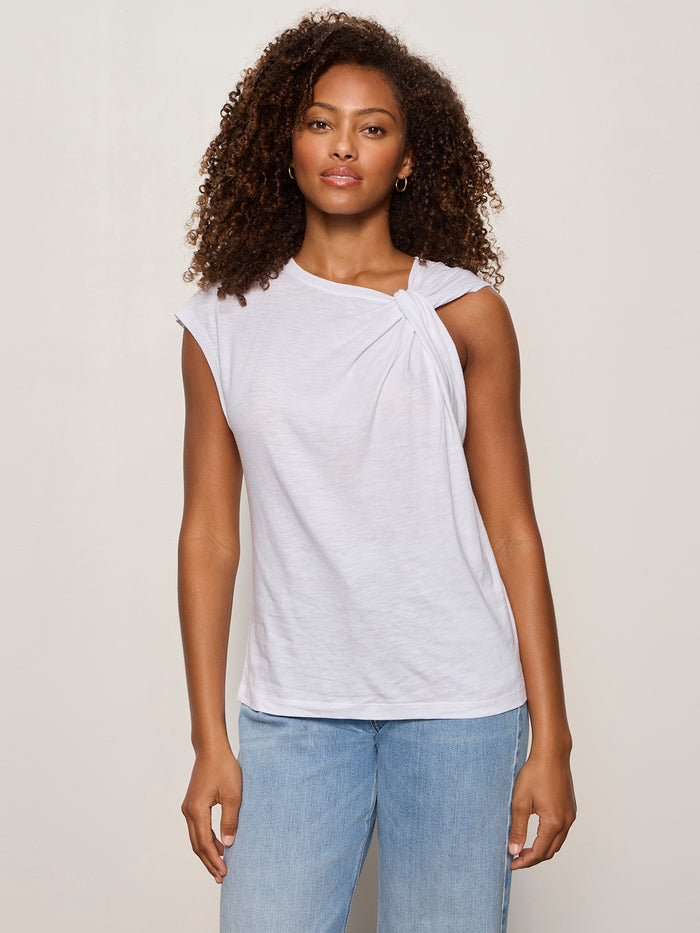 A woman with curly hair wears the Sanctuary Clothing “so twisted muscle tee” in white and light blue jeans, standing against a plain off-white background.