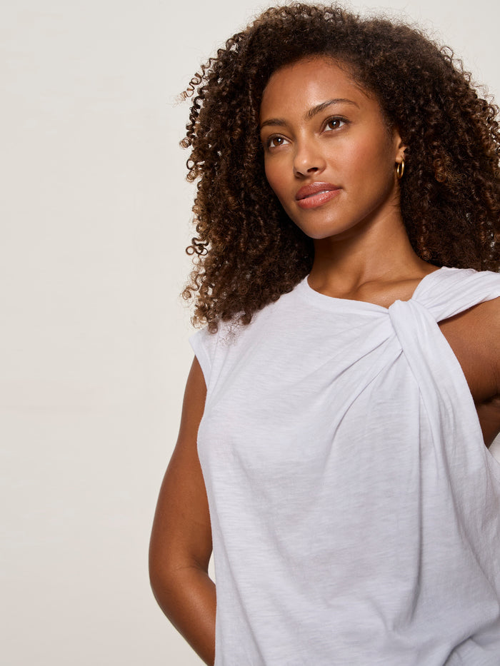 A woman with curly hair wears the Sanctuary Clothing "so twisted muscle tee" in white, featuring a knotted one-shoulder detail, standing against a plain light background and gazing calmly to the side.