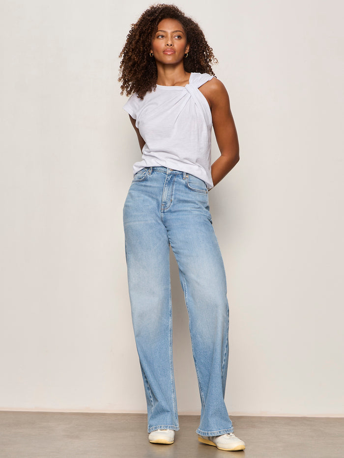 A woman with curly hair stands against a light wall, wearing the Sanctuary Clothing so twisted muscle tee in white and light blue high-waisted wide-leg jeans, paired with cream-colored shoes.