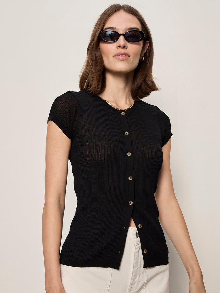 A woman wearing the Sanctuary Clothing soft focus ribbed top in black and light pants stands against a plain light background, looking slightly to the side, accessorized with black sunglasses.