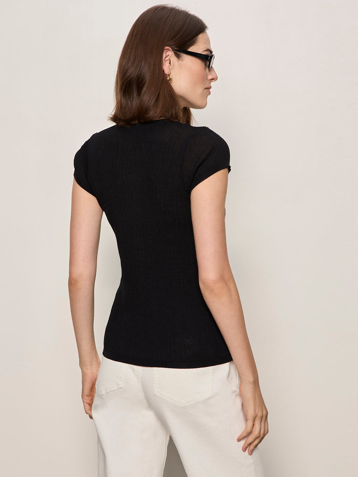 A woman with straight brown hair wears black sunglasses, white pants, and the Sanctuary Clothing soft focus ribbed top in black, standing with her back to a light-colored wall.