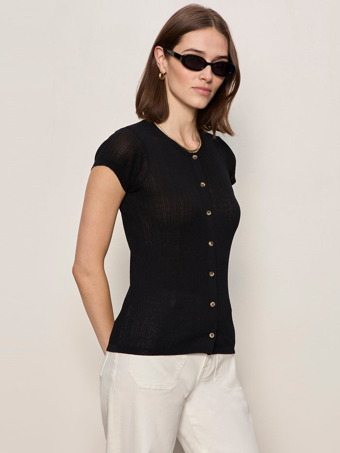 A woman wears the Sanctuary Clothing Soft Focus Ribbed Top in black, styled with light pants, gold hoop earrings, and black sunglasses, standing against a light background with her hands behind her back.