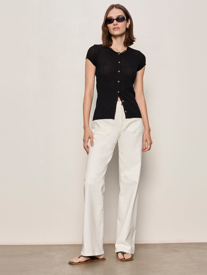 A woman with shoulder-length brown hair and a relaxed posture stands against a light wall, wearing black sunglasses, white wide-leg pants, sandals, and the Sanctuary Clothing soft focus ribbed top in black.
