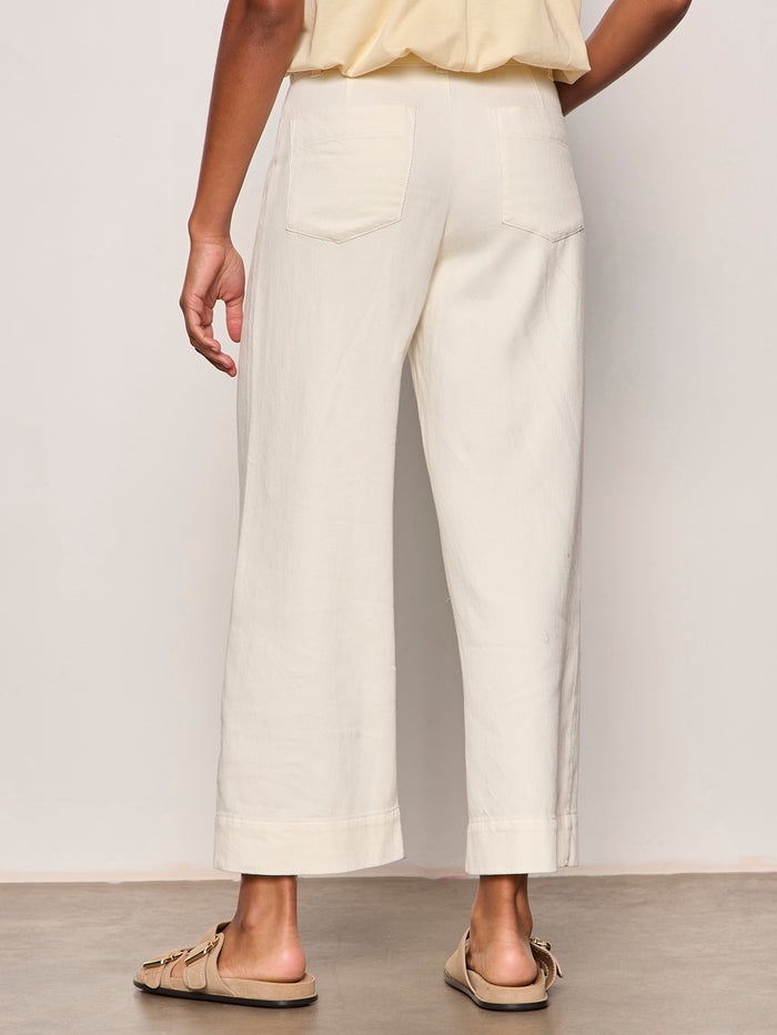 A person faces away, wearing Sanctuary Clothing's soft marine wide leg crop pant collagen in off-white with back pockets, paired with a cream top and beige slide sandals, standing on a light floor against a plain background.