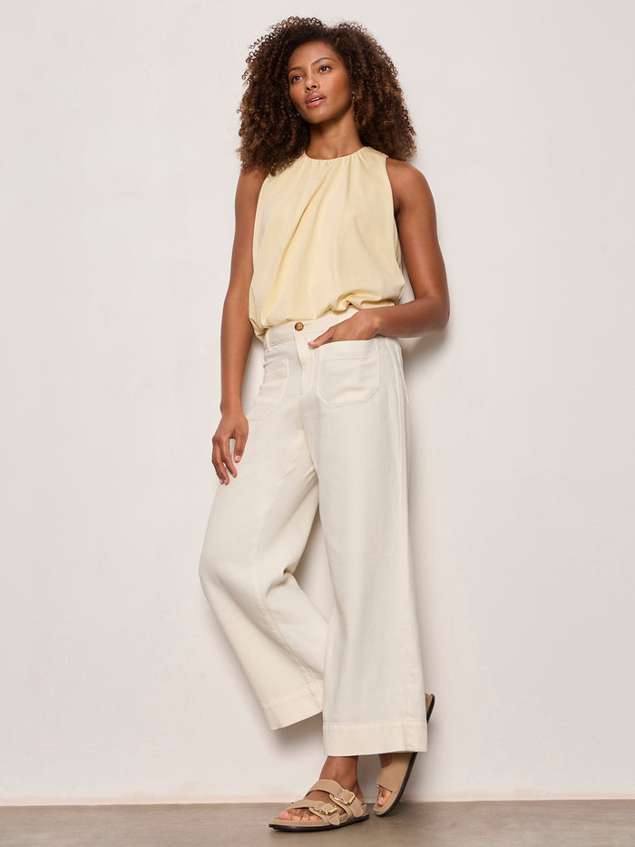 A woman with curly hair stands against a plain wall, wearing Sanctuary Clothing's soft marine wide leg crop pant collagen, paired with a sleeveless light yellow top and beige sandals. She has one hand in her pocket and the other by her side.