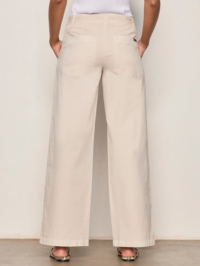 Wearing Sanctuary Clothing’s soft polished wide leg summer sand pants, featuring large back pockets, a person stands against a light wall with hands in their pockets; only the lower back half is visible.