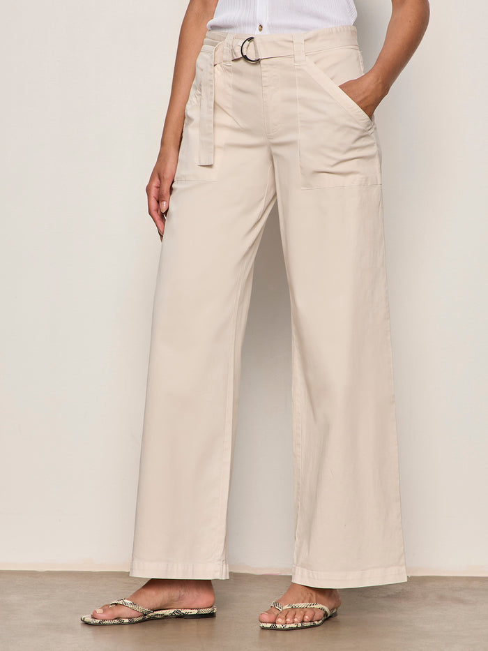 A person wearing Sanctuary Clothing's soft polished wide leg summer sand trousers, featuring large front pockets and a matching belt, paired with a tucked-in white top and snakeskin flip-flops, stands against a plain background.