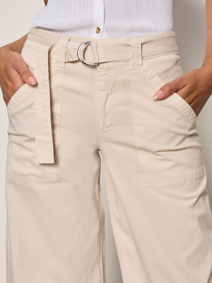A close-up of a person wearing Sanctuary Clothing's soft polished wide leg summer sand pants with a fabric belt and silver buckle, paired with a white sleeveless top. The person's hands are tucked into the front pockets.
