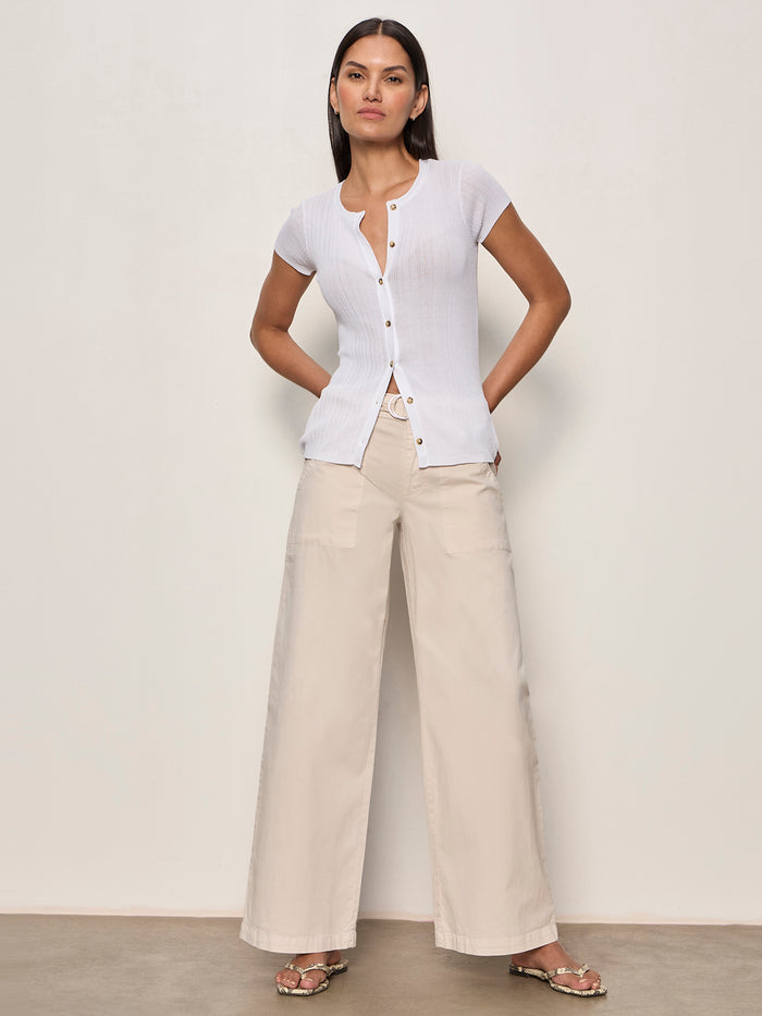 A woman with straight dark hair and a confident expression stands against a plain wall, wearing Sanctuary Clothing's soft polished wide leg summer sand pants, paired with a short-sleeve white button-up top and sandals.