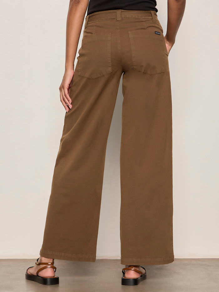 A woman is seen from the back wearing Sanctuary Clothing's soft polished wide leg timber pants and black sandals, with her right hand on her hip, standing against a plain light wall.