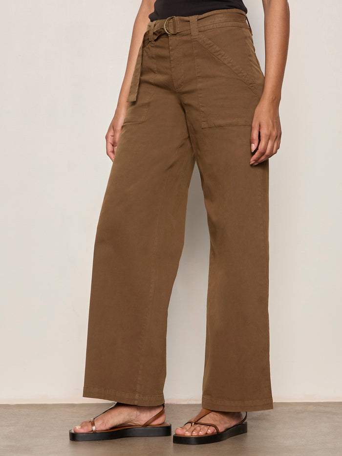 A person wearing Sanctuary Clothing's "soft polished wide leg timber" high-waisted brown pants with large front pockets and a matching belt, paired with black sandals, stands against a light-colored wall.