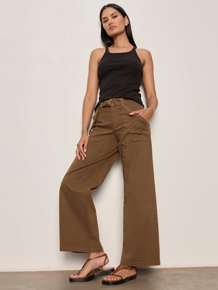 A woman with straight dark hair, one hand in her pocket, wears Sanctuary Clothing's soft polished wide leg timber pants with a belt, a black tank top, and black sandals against a plain light-colored wall.