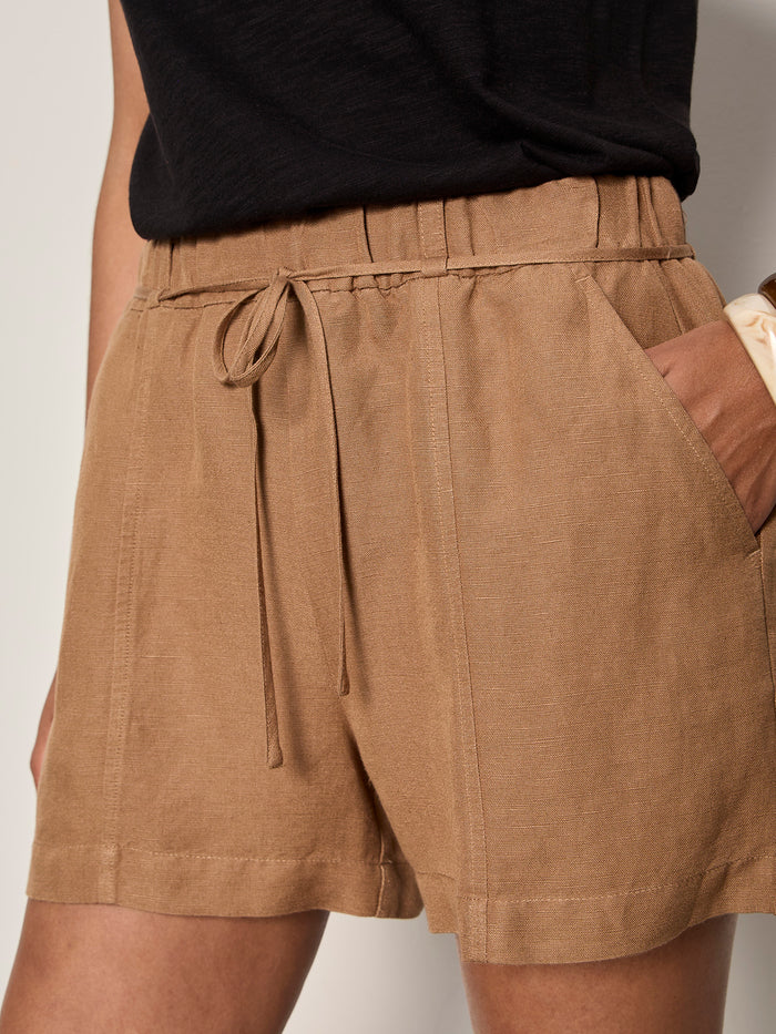 A person wears Sanctuary Clothing’s springtide short in toasted coconut—a high-waisted, relaxed fit short with visible stitching, side pockets, and a drawstring—paired with a black top and one hand in the pocket.