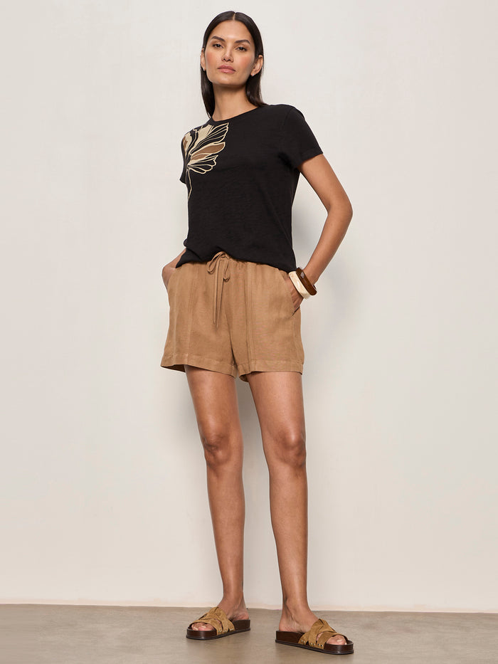 A woman stands against a plain light wall wearing Sanctuary Clothing springtide short in toasted coconut, a black T-shirt with gold leaf design, brown sandals, and bracelets. Her hands are in her pockets as she looks at the camera.