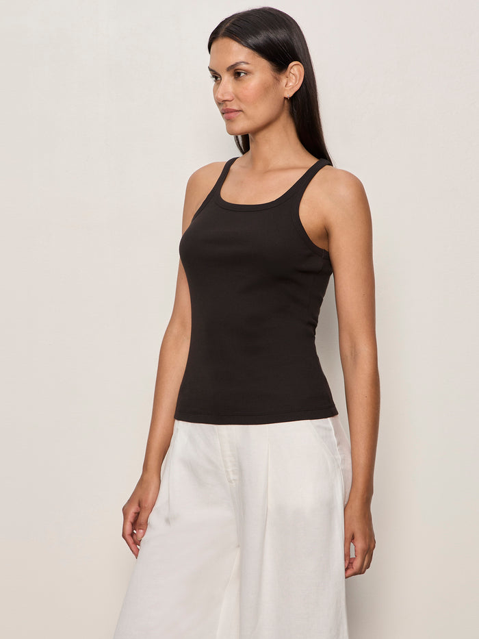 A woman with long dark hair wears the Sanctuary Clothing sprinter rib tank in black, paired with loose white pants. She stands against a plain light wall, looking slightly to the side.