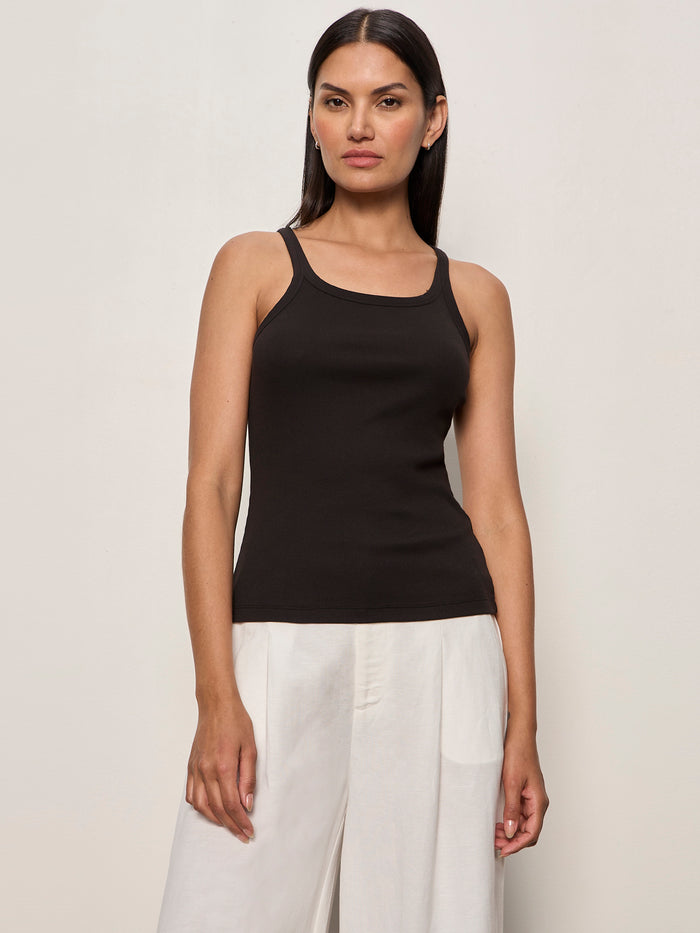 A woman with long dark hair wears the Sanctuary Clothing sprinter rib tank in black, paired with loose white pants, standing against a plain light background and facing forward with a neutral expression.