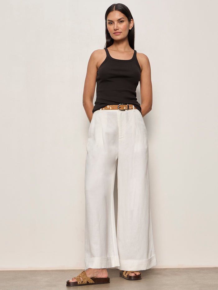 A woman with straight dark hair stands against a plain wall, wearing the Sanctuary Clothing sprinter rib tank in black. She pairs it with white wide-legged trousers, a brown belt, and brown sandals, her hands tucked in her pockets.