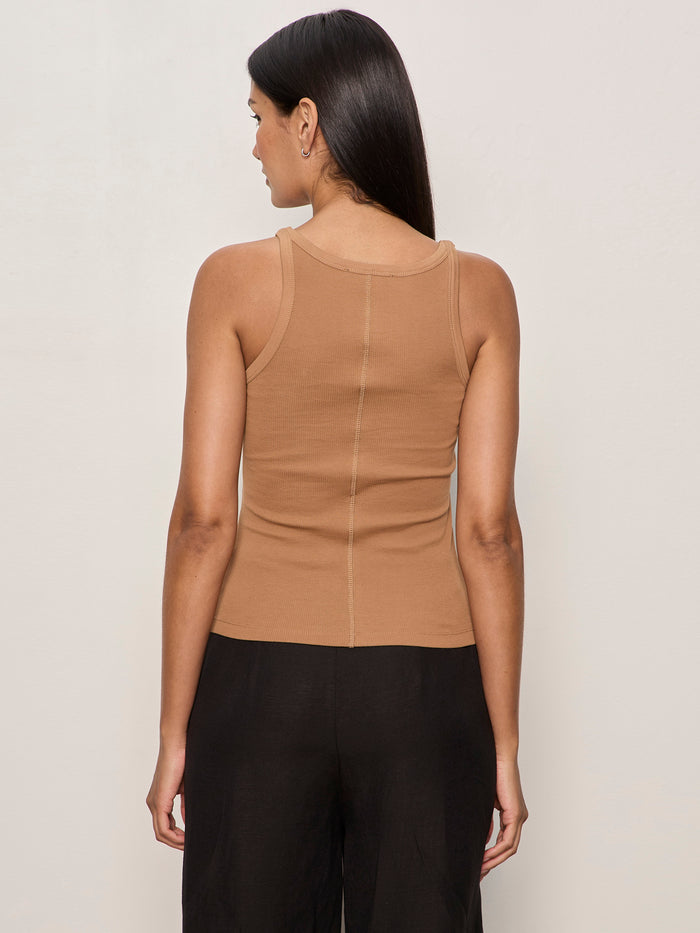 A woman with long dark hair faces away, wearing the Sanctuary Clothing sprinter rib tank in toasted coconut and black pants, set against a plain light-colored background.
