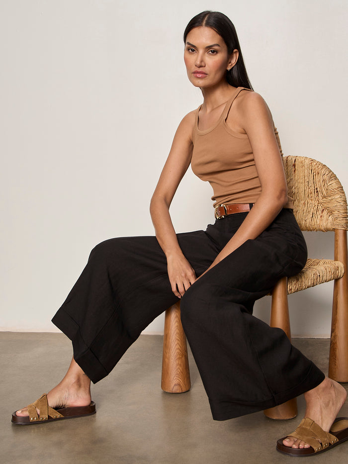 A woman with long dark hair sits on a woven chair wearing the Sanctuary Clothing sprinter rib tank in toasted coconut, paired with black wide-leg pants, a brown belt, and brown sandals. She gazes neutrally at the camera against a minimal background.