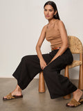 A woman with long dark hair and a neutral expression sits on a woven chair, wearing Sanctuary Clothing's terrace linen pant in black, a tan sleeveless top, brown belt, and tan fringe slide sandals. The background is plain.