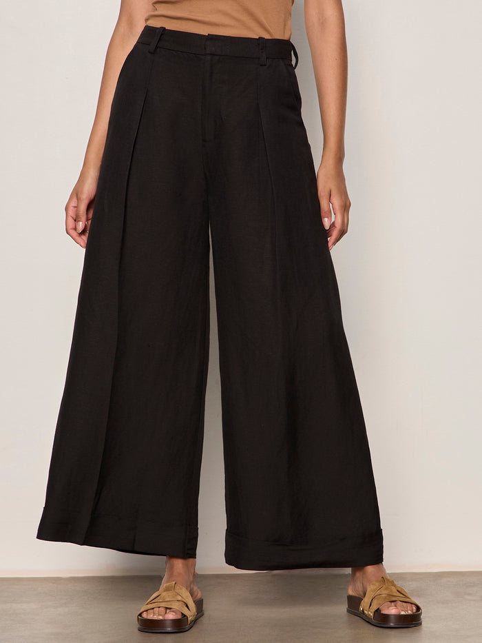 Wearing Sanctuary Clothing's terrace linen pant in black, paired with a tan top and tan sandals, a person stands against a light wall with arms at their sides. Photo is cropped from shoulders to feet.