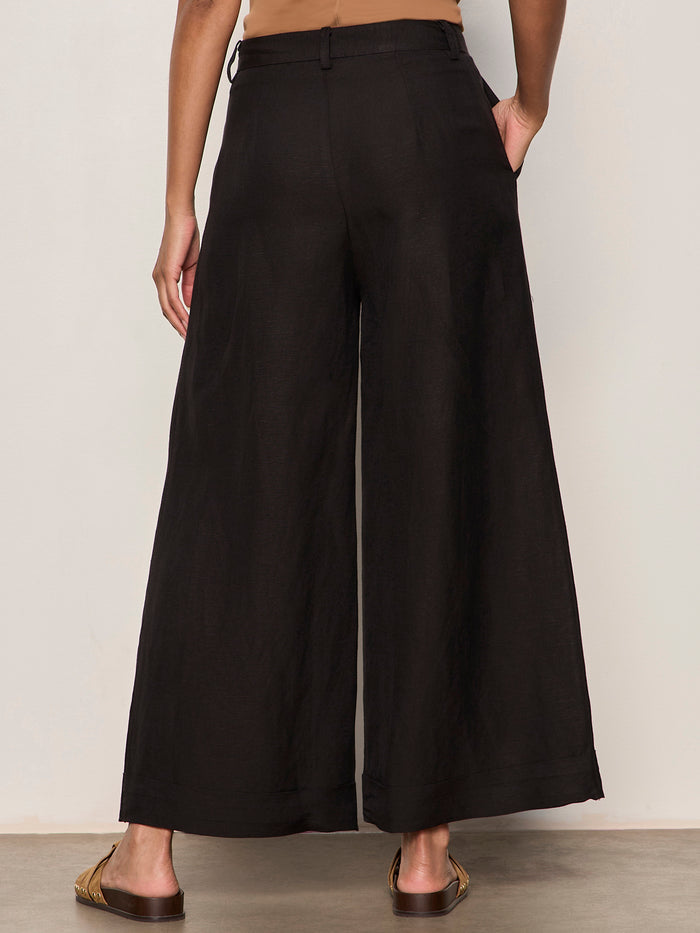 A person stands facing away, wearing Sanctuary Clothing's terrace linen pant in black with brown sandals, left hand in their pocket against a plain, light-colored wall.